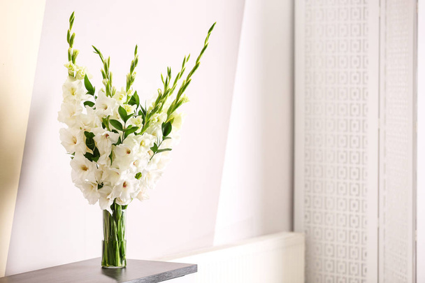 Vase with beautiful white gladiolus flowers on wooden table near color wall. Space for text - Foto, imagen