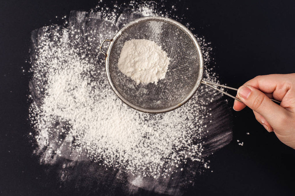 white flour and strainer in hand on the black background - Foto, Imagen