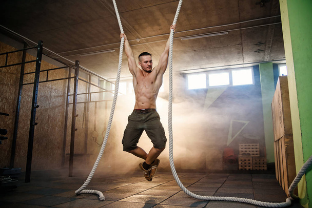 Strong young crossfitter climbing on the rope during training. - Фото, изображение
