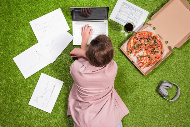 Woman having lunch with pizza and glass of wine working  on the grass using laptop with graphics and charts printed on the paper. - Φωτογραφία, εικόνα
