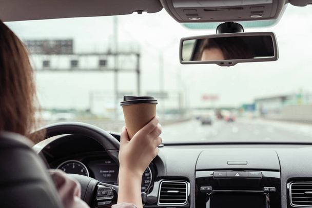 woman is driving on the highway and holding paper cup of coffee at the morning . View from the back seat of the car  - Zdjęcie, obraz