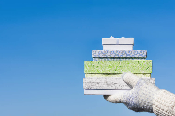 Woman hand in the knitted gloves with many gift boxes on the blue sky background. Concept of seasonal or winter discount - Foto, afbeelding