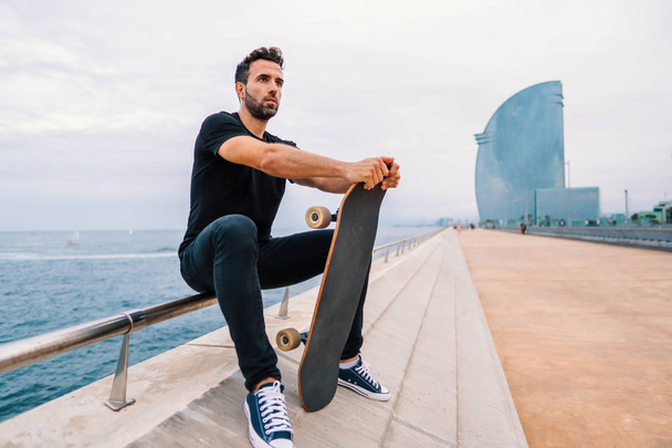 Skateboarder siedzi na deskorolce na nowoczesnym miejskim tarasie morskim - Zdjęcie, obraz