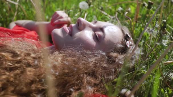 カーリー魅力的な女の子は緑の草の上に横たわっています。若いかわいい女性が目を閉じた。赤いTシャツを着た陽気な女性。夏休みのコンセプト. - 映像、動画