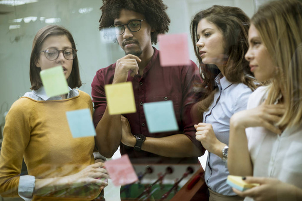 Група молодих бізнесменів обговорює перед скляною стіною, використовуючи нотатки та наклейки на стартовому офісі
 - Фото, зображення