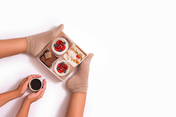 Creative photo of female legs  with a cup of coffee and a box of small cakes on a white background. Concept. Top view  - Photo, Image