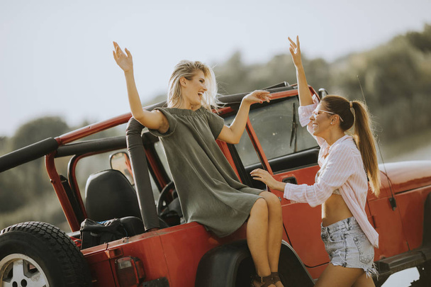 Две молодые женщины веселятся на капоте машины у озера в жаркий летний день
 - Фото, изображение