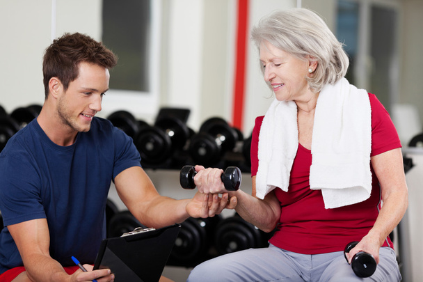 Instructor Assisting Senior Woman In Lifting Dumbbells At Gym - 写真・画像