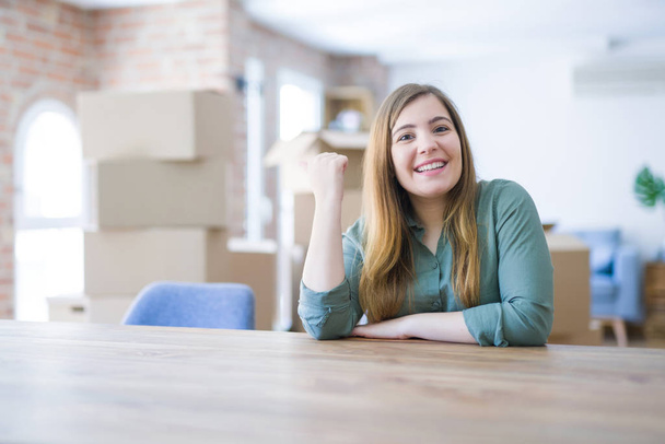 Mujer joven sentada en la mesa con cajas de cartón detrás de su mudanza a un nuevo hogar sonriendo con la cara feliz mirando y señalando a un lado con el pulgar hacia arriba
. - Foto, imagen