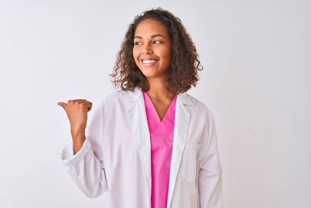 Młody brazylijski lekarz kobieta noszenie płaszcz stojący na białym tle uśmiechnięty z szczęśliwy twarz patrząc i wskazując na bok z kciukiem w górę. - Zdjęcie, obraz