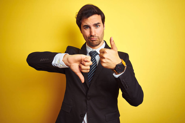 Young handsome businessman wearing suit and tie standing over isolated yellow background Doing thumbs up and down, disagreement and agreement expression. Crazy conflict - Photo, Image