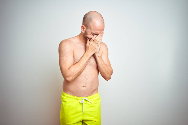 Молодой человек без рубашки в отпуске в жёлтых купальниках на изолированном фоне с грустным лицом, прикрывающим лицо руками во время плача. Концепция депрессии
. - Фото, изображение
