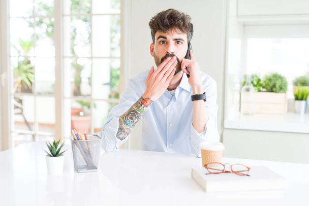 junger Geschäftsmann spricht auf Smartphone-Cover Mund mit Hand schockiert vor Scham für Fehler, Ausdruck der Angst, Angst in der Stille, geheimes Konzept - Foto, Bild