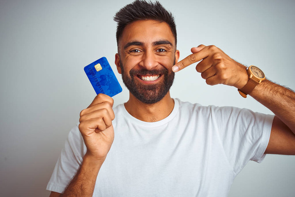 Nuori intialainen asiakas mies tilalla luottokortti seisoo yli eristetty valkoinen tausta erittäin onnellinen osoittaa käsin ja sormella
 - Valokuva, kuva