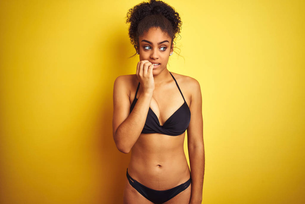 Afroamerikai nő a nyaralás visel bikini alatt álló elszigetelt sárga háttér látszó hangsúlyozta és ideges kézzel a száj harapós körmök. Szorongás probléma. - Fotó, kép
