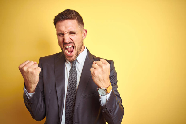 Young handsome business man over yellow isolated background very happy and excited doing winner gesture with arms raised, smiling and screaming for success. Celebration concept. - Photo, image