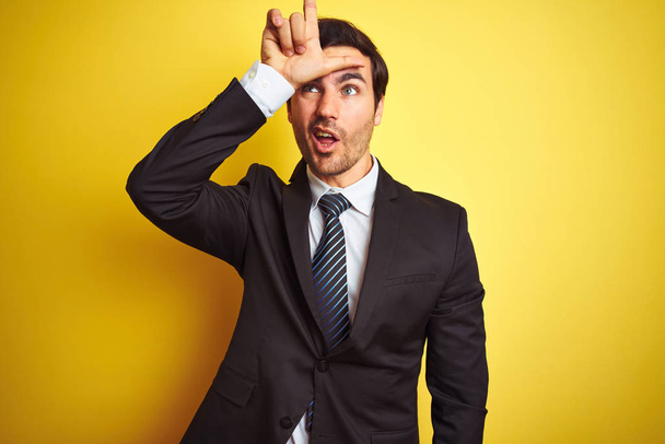 Young handsome businessman wearing suit and tie standing over isolated yellow background making fun of people with fingers on forehead doing loser gesture mocking and insulting. - Photo, Image