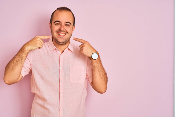 Nuori mies yllään tyylikäs paita seisoo yli eristetty vaaleanpunainen tausta hymyilevä iloinen osoittaa ja sormilla hampaat ja suu. Hammasterveyden käsite
. - Valokuva, kuva