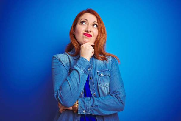 Young beautiful redhead woman wearing denim shirt standing over blue isolated background Thinking worried about a question, concerned and nervous with hand on chin - Foto, Bild