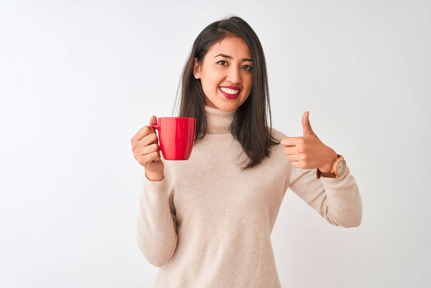 Beautiful chinese woman drinking red cup of coffee standing over isolated white background happy with big smile doing ok sign, thumb up with fingers, excellent sign - Zdjęcie, obraz