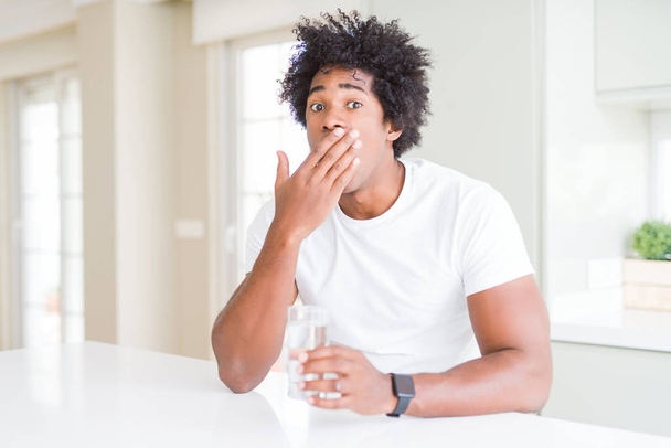 African American man die het drinken van een glas water thuis cover mond met de hand met schaamte voor fout, uitdrukking van angst geschokt, bang in stilte, geheime concept - Foto, afbeelding