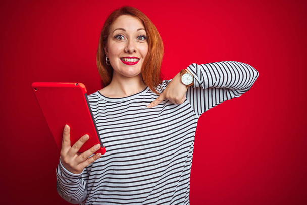 Jonge mooie roodharige vrouw met behulp van Tablet over rode geïsoleerde achtergrond met verrassing gezicht wijzende vinger naar zichzelf - Foto, afbeelding
