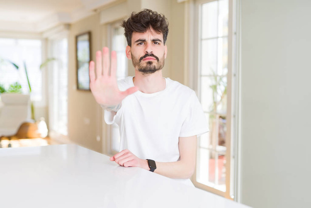 Jonge man dragen casual t-shirt zittend op witte tafel doen stoppen met het zingen met Palm van de hand. Waarschuwings uitdrukking met negatief en serieus gebaar op het gezicht. - Foto, afbeelding