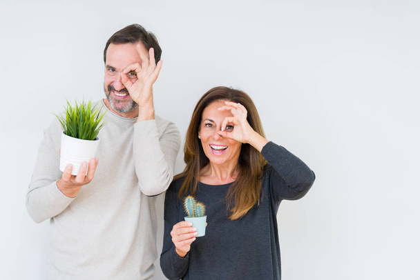 Middle age couple holding plants over isolated background with happy face smiling doing ok sign with hand on eye looking through fingers - 写真・画像