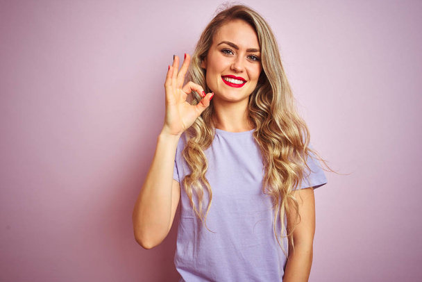 Joven hermosa mujer vistiendo púrpura camiseta de pie sobre rosa aislado fondo sonriendo positivo haciendo signo de ok con la mano y los dedos. Expresión exitosa
. - Foto, Imagen