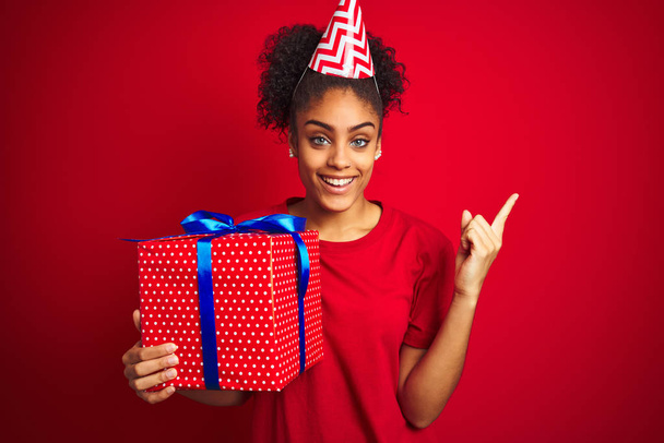 donna afroamericana indossa cappello divertente tenendo regalo di compleanno su sfondo rosso isolato molto felice puntando con mano e dito di lato
 - Foto, immagini