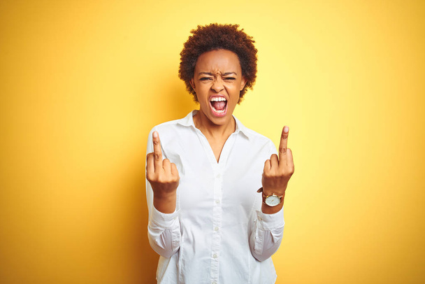 Afrika kökenli Amerikalı iş kadını izole edilmiş sarı arka planda orta parmağını göstererek kötü ifade, provokasyon ve kaba tavır sergiliyor. Heyecanlı çığlık - Fotoğraf, Görsel