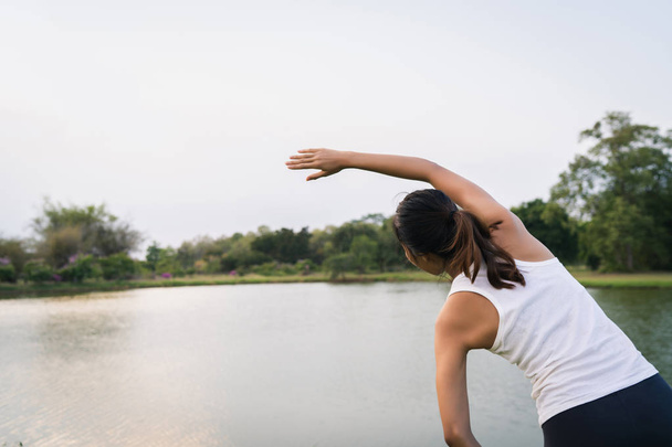 Terve nuori aasialainen juoksija nainen lämmittää kehoa venyttämällä ennen liikuntaa ja joogaa lähellä järveä puistossa lämpimän valoisan aamun alla. Elämäntapa kunto ja aktiiviset naiset liikunta kaupunkien konseptissa
. - Valokuva, kuva