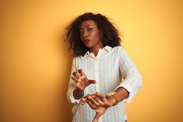 孤立した黄色の背景に立つストライプのシャツを着たアフリカ系アメリカ人女性は、嫌悪感を抱くため嫌な表情を浮かべた。手を上げて迷惑な概念. - 写真・画像