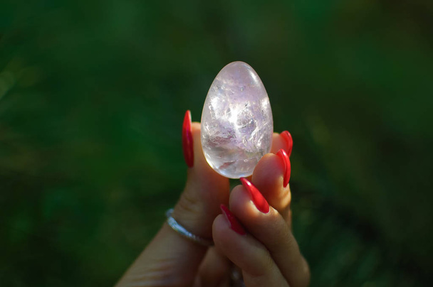 Female hand with red manicure holding transparent violet amethyst yoni egg for vumfit, imbuilding or meditation. Crystal quartz egg in hands on green background outdoors - Fotoğraf, Görsel