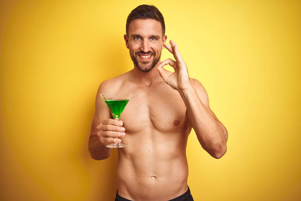 Mladý pohledný muž, který pije Letní koktejl přes izolované žluté pozadí, se dobře podepíší prsty, skvělý symbol - Fotografie, Obrázek