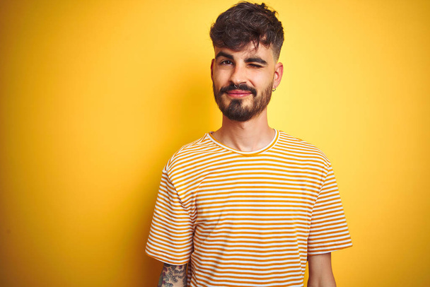 Mladý muž s tetováním v proužkované košili, stojící nad izolovaným žlutým pozadím, se dívá na fotoaparát s sexy výrazem, veselou a šťastnou tváří. - Fotografie, Obrázek