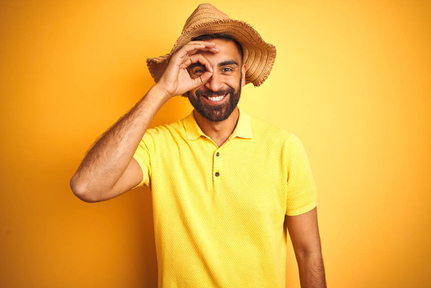 Jovem indiano de férias usando chapéu de verão em pé sobre fundo amarelo isolado fazendo gesto ok com a mão sorrindo, olho olhando através dos dedos com rosto feliz
. - Foto, Imagem