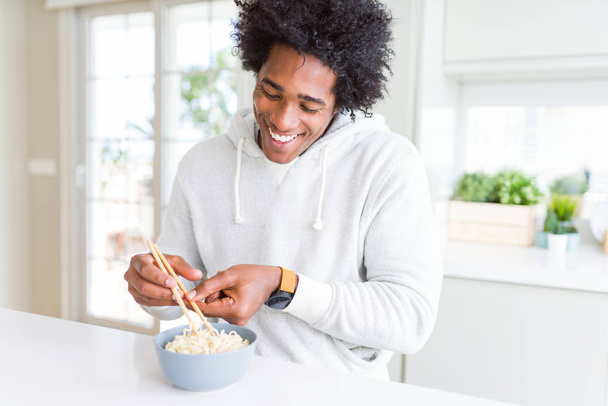Afro-amerikai ember eszik ázsiai tészta segítségével pálcika otthon egy boldog arc állt, és mosolyogva egy magabiztos mosoly mutatja fogak - Fotó, kép