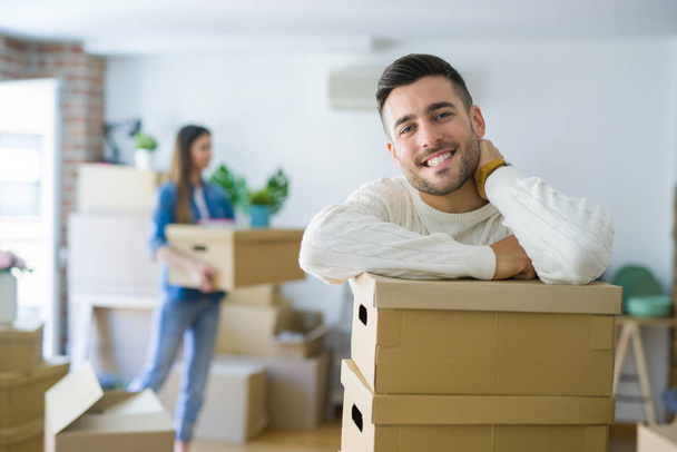 Young couple moving to a new home, handsome man smiling leaning on cardboard boxes - Photo, Image