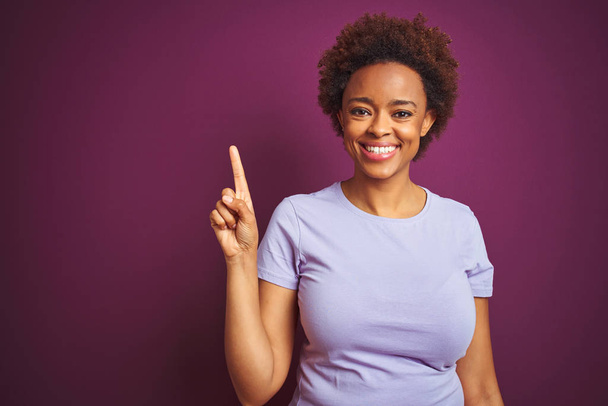 孤立した紫色の背景の上にアフロの髪を持つ若い美しいアフリカ系アメリカ人女性は、自信を持って幸せに微笑みながら、指のナンバーワンで見せ、指を上げる. - 写真・画像
