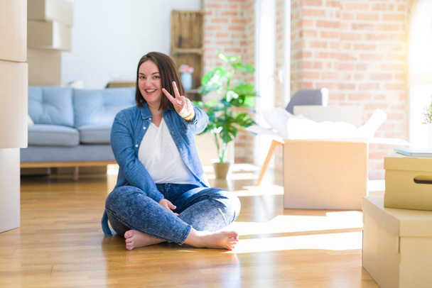 Young plus size woman sitting on the floor around cardboard boxes moving to a new home smiling with happy face winking at the camera doing victory sign. Number two. - Foto, immagini