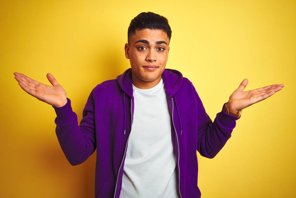 孤立した黄色の背景の上に立つ紫色のスウェットシャツを着た若いブラジル人男性は、腕と手を上げて混乱した表情を浮かべた。疑いの概念. - 写真・画像