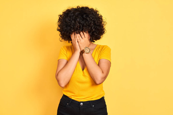 Νέα αραβική γυναίκα με σγουρά μαλλιά φορώντας t-shirt στέκεται πάνω από απομονωμένο κίτρινο φόντο με θλιβερή έκφραση καλύπτοντας το πρόσωπο με τα χέρια ενώ κλαίει. Έννοια της κατάθλιψης. - Φωτογραφία, εικόνα