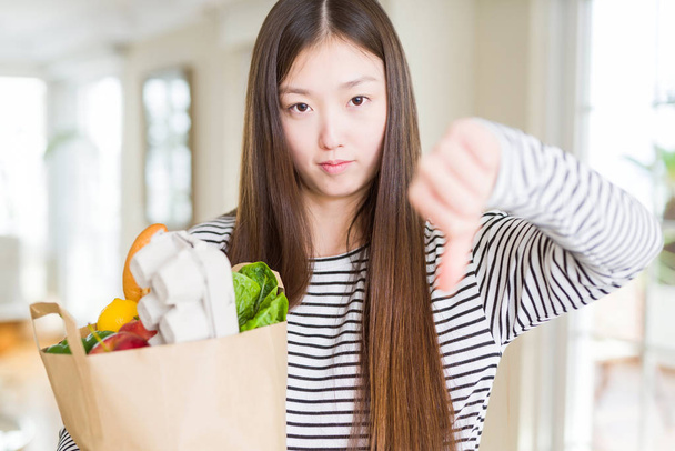 Hermosa mujer asiática sosteniendo bolsa de papel de alimentos frescos con la cara enojada, signo negativo que muestra aversión con los pulgares hacia abajo, concepto de rechazo
 - Foto, Imagen
