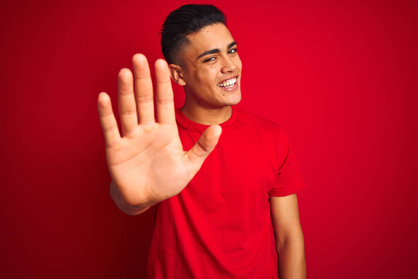 Jovem brasileiro vestindo camiseta em pé sobre fundo vermelho isolado Waiving dizendo olá feliz e sorridente, gesto de boas-vindas amigável
 - Foto, Imagem