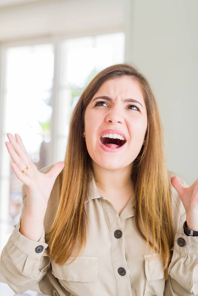 Beautiful young woman at home crazy and mad shouting and yelling with aggressive expression and arms raised. Frustration concept. - Фото, изображение