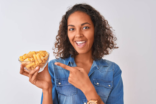 Young brazilian woman holding bowl with macaroni pasta over isolated white background very happy pointing with hand and finger - Fotó, kép