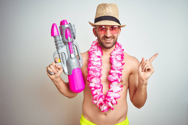 Young man wearing summer hat and hawaiian lei flowers holding water gun over isolated background very happy pointing with hand and finger to the side - Photo, Image
