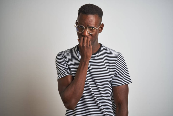 Uomo afroamericano indossa maglietta a righe e occhiali su sfondo bianco isolato cercando stressato e nervoso con le mani in bocca mordere le unghie. Problema di ansia
. - Foto, immagini