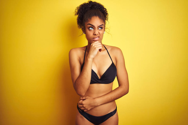 Afrikanerin im Urlaub im Bikini vor isoliertem gelben Hintergrund, besorgt über eine Frage, besorgt und nervös mit der Hand am Kinn - Foto, Bild